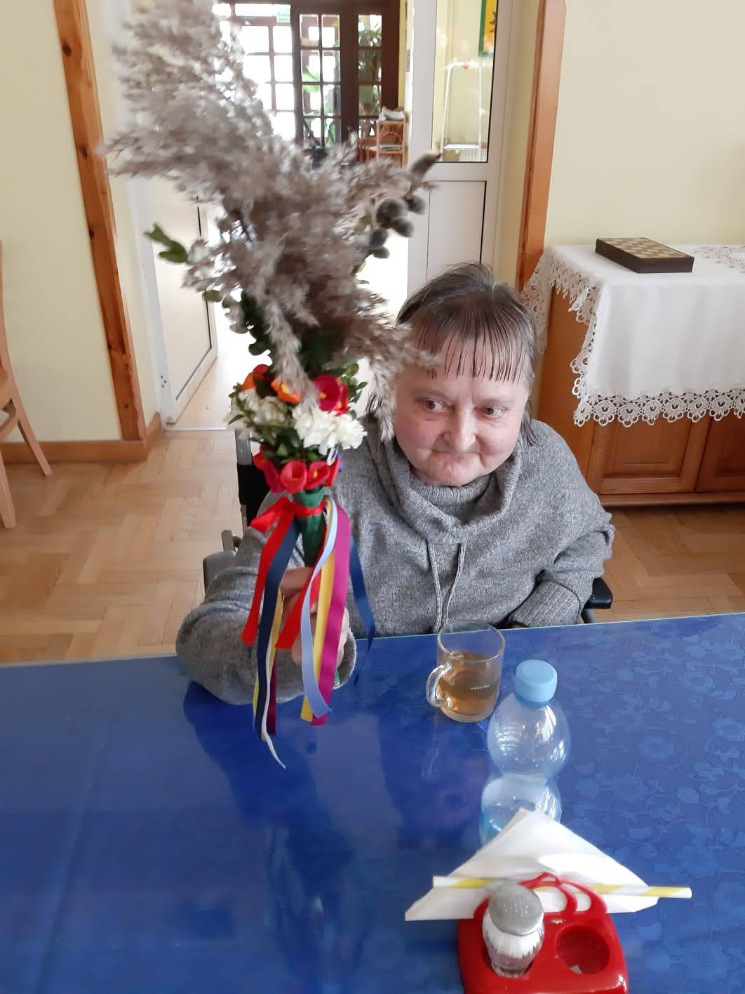 Kobieta siedząca przy stole trzyma palmę, kobieta lekko się uśmiecha.