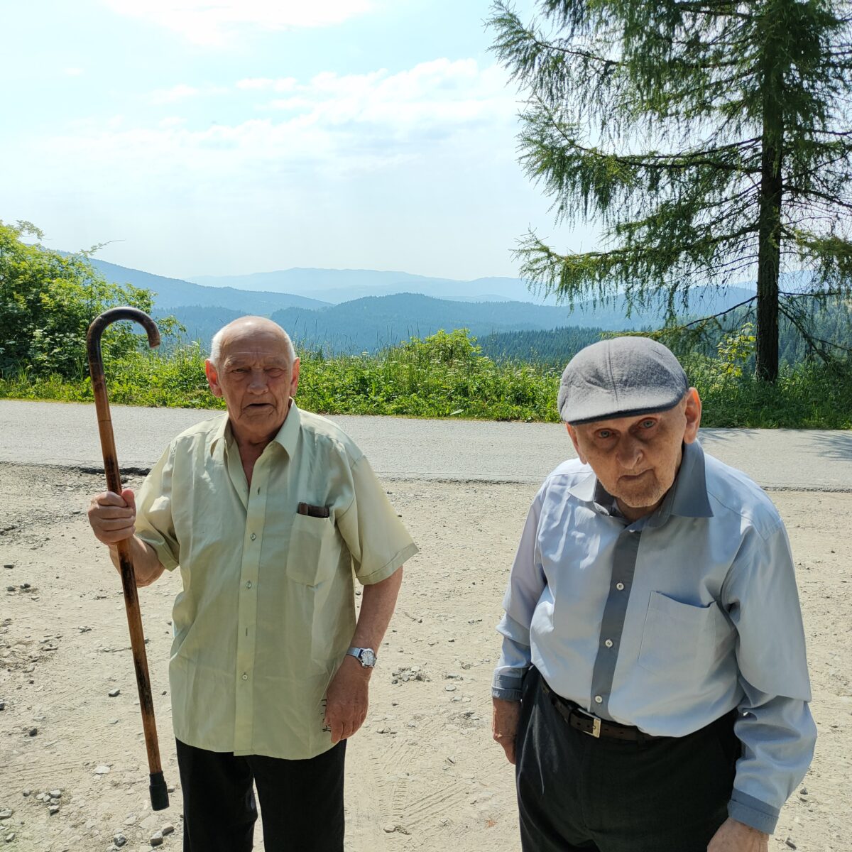Dwóch starszych mężczyzn stoi i patrzy w obiektyw. Jeden z nich trzyma w ręku laskę, którą zaczepnie unosi w górę, drugi ma na głowie kaszkiet. W tle widać ulicę a dalej zielone krzewy i góry.
