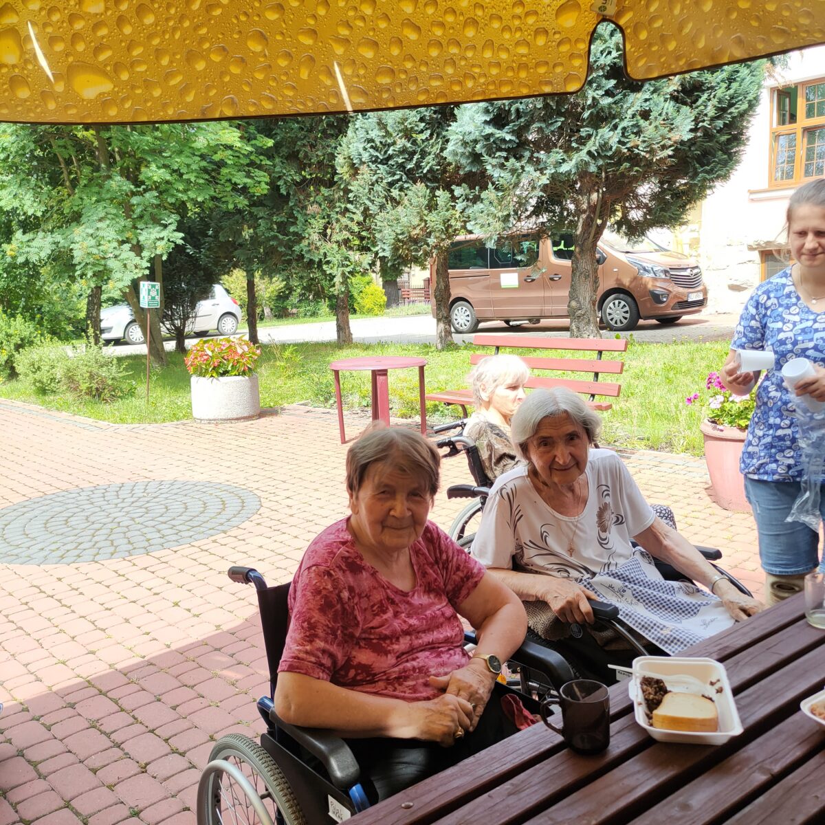 Dwie starsze kobiety na wózkach inwalidzkich, siedzą przy stole, uśmiechają się do obiektywu, na stole jest kubek i talerzyk z chlebem, po prawej stronie kobieta niesie dwa kubki z wodą