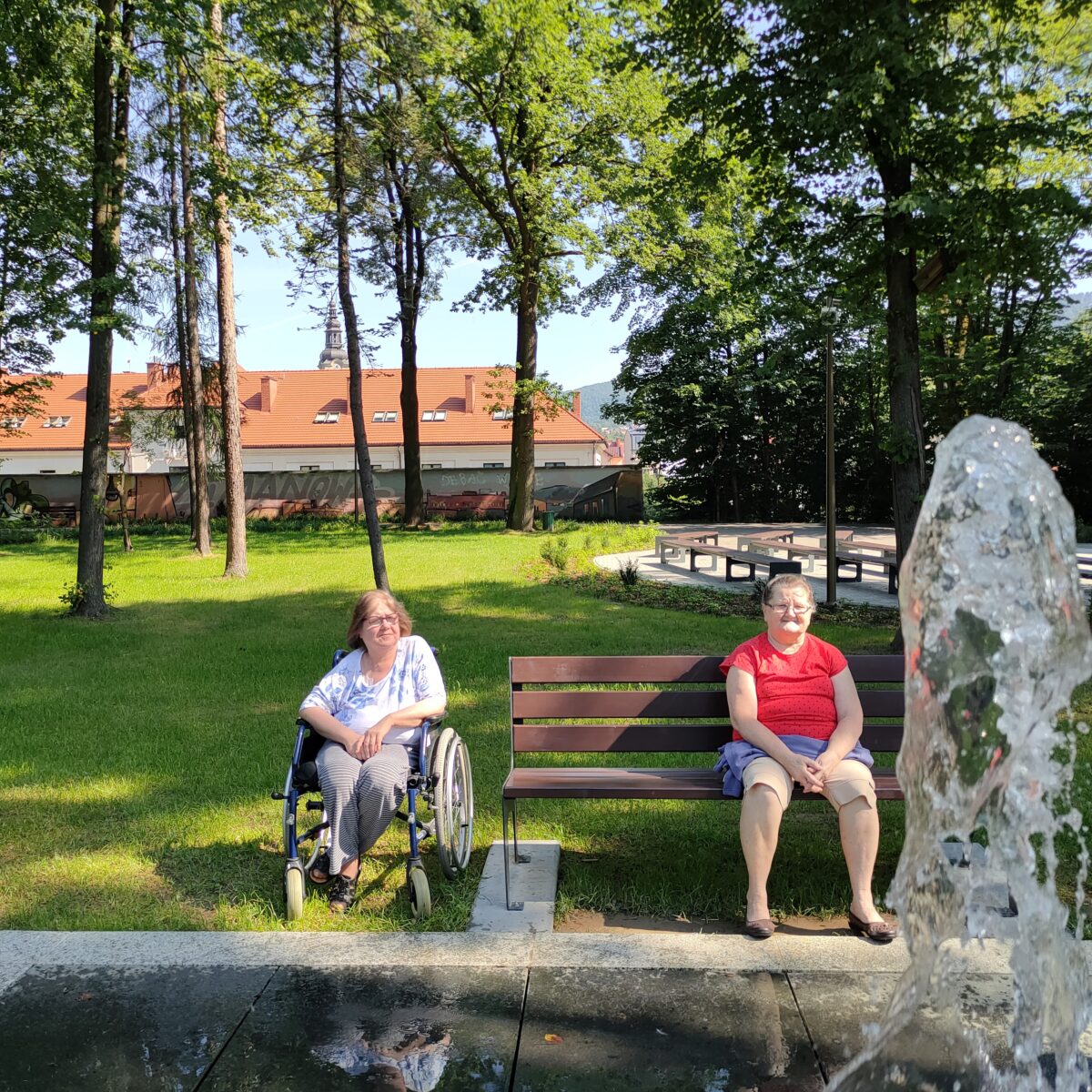 Na ławce siedzi starsza kobieta w czerwonej koszulce, obok ławki na wózku inwalidzkim siedzi druga kobieta. Na pierwszym planie widać wodę z fontanny, w tle drzewa i dach budynku.