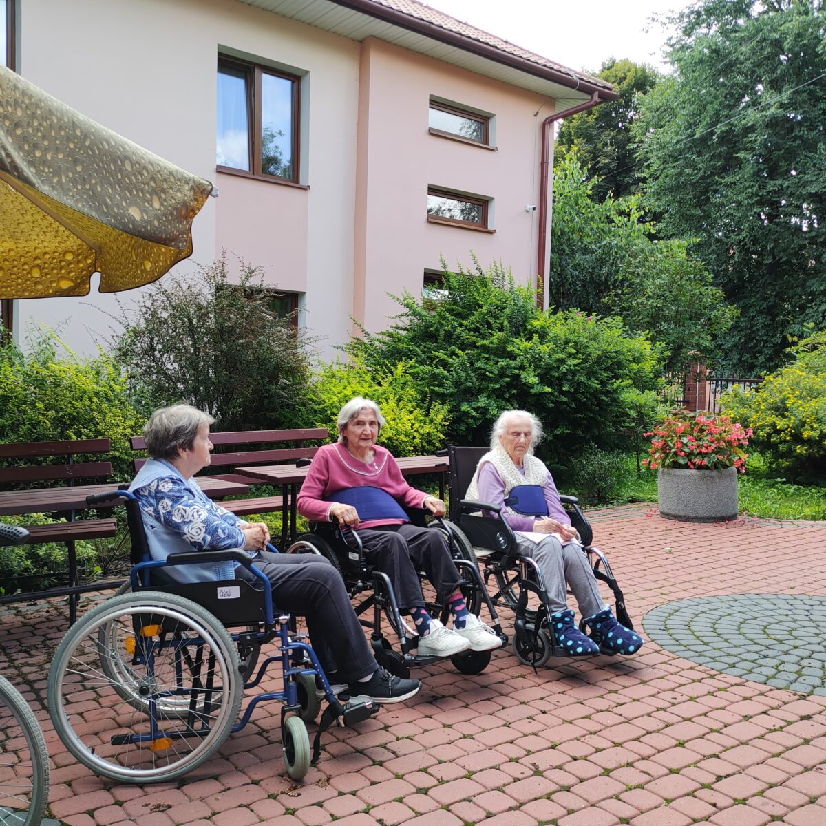 Trzy starsze kobiety siedzące na wózkach inwalidzkich. W tle kawałek żółtego dużego parasola przeciwsłonecznego oraz kawałek budynku. Widać również zielone krzewy i drzewa. Na ziemi jest kostka brukowa w kolorze bordowym i szarym.
