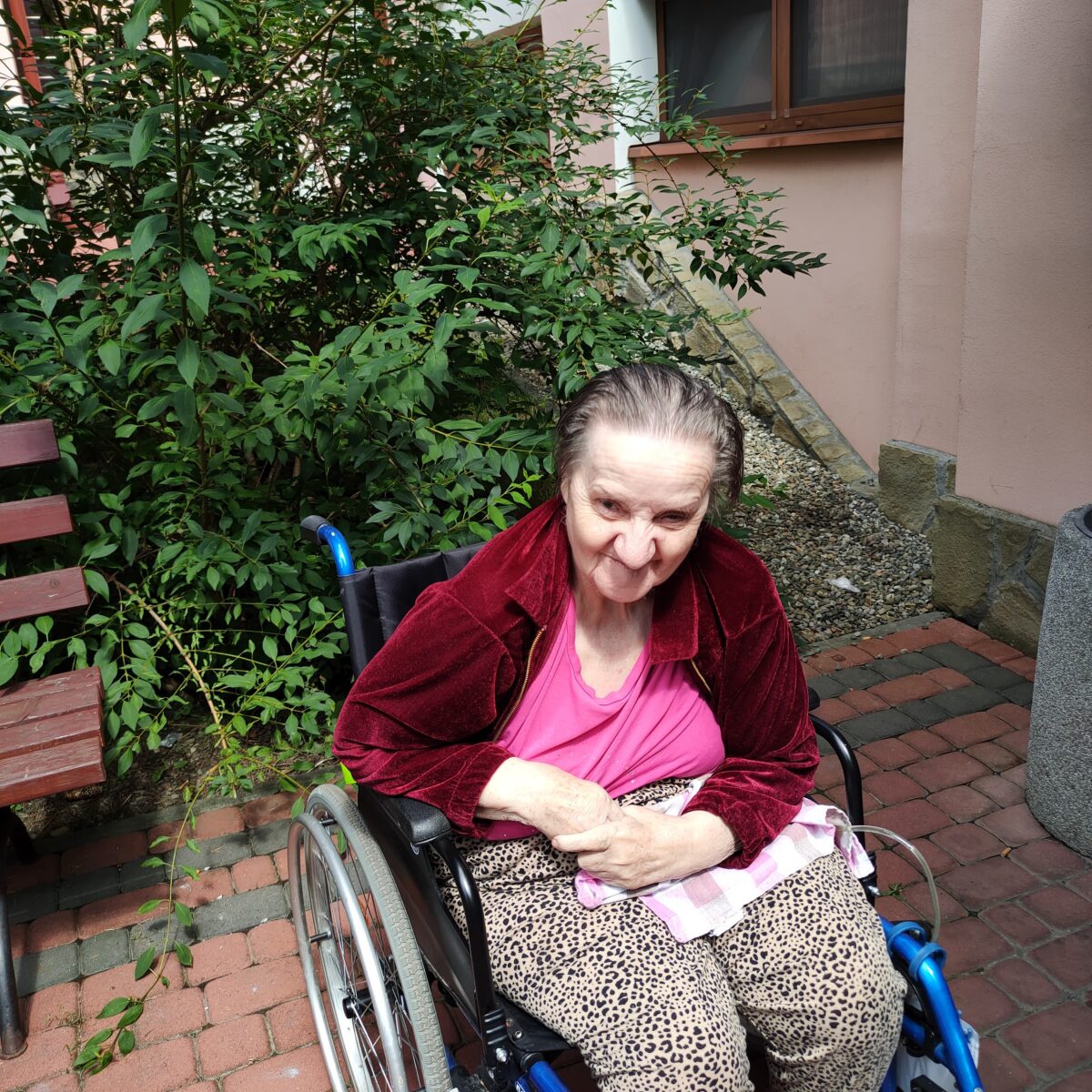 Kobieta na wózku inwalidzkim uśmiecha się, za nią widać zielony krzew i kawałek budynku.