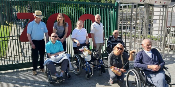 Na tle bramy z napisem "ZOO" stoją mężczyźni i kobiety, w pierwszym rzędzie dwie osoby na wózkach inwalidzkich, po prawej stronie mężczyzna na wózku inwalidzkim a za nim kucająca kobieta w okularach przeciwsłonecznych. Zdjęcie jest jasne.