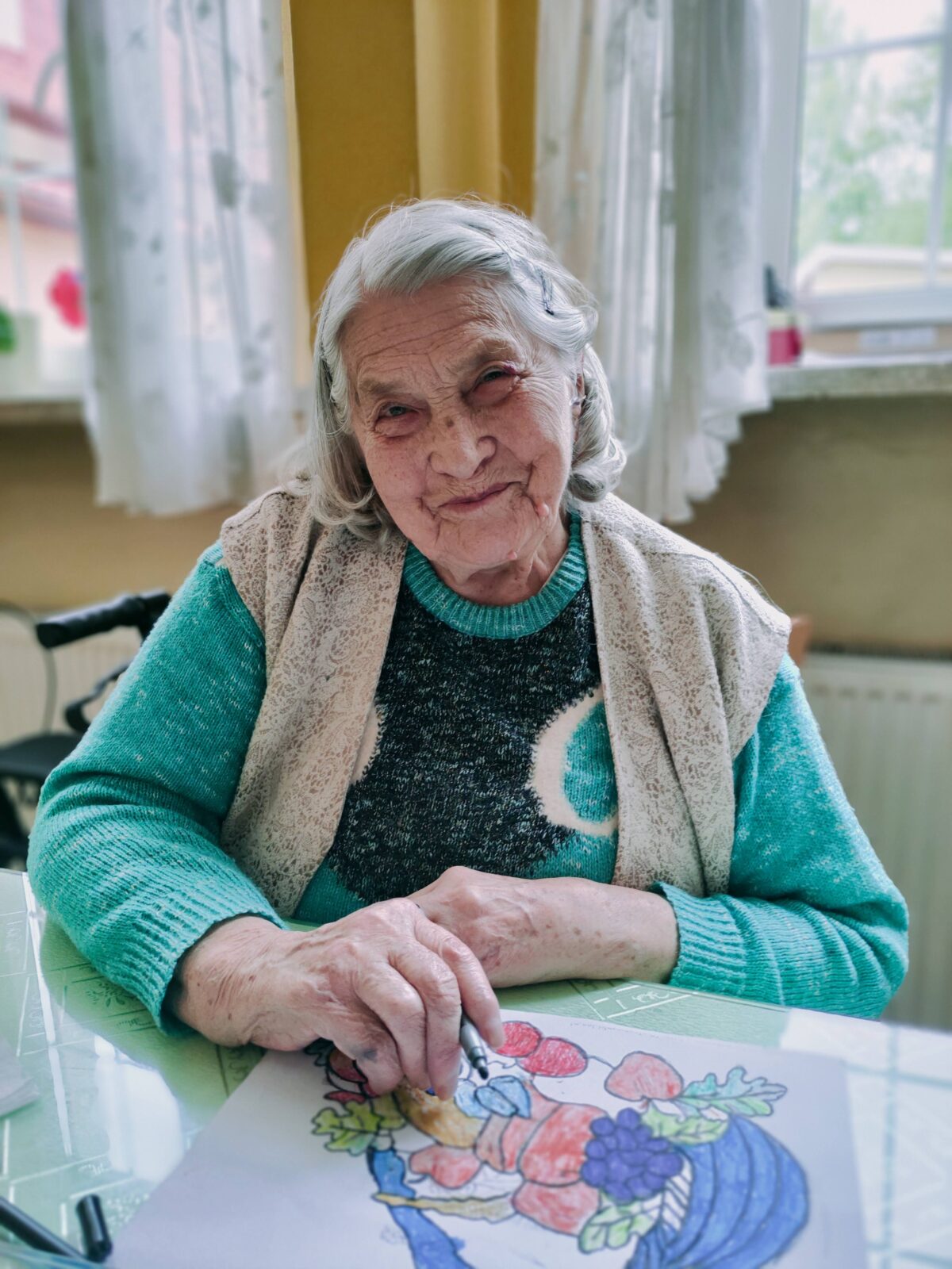 Uśmiechnięta starsza kobieta siedząca przy stole. W ręku ma kredkę, na stole jest rysunek. Kobieta patrzy prosto w obiektyw.