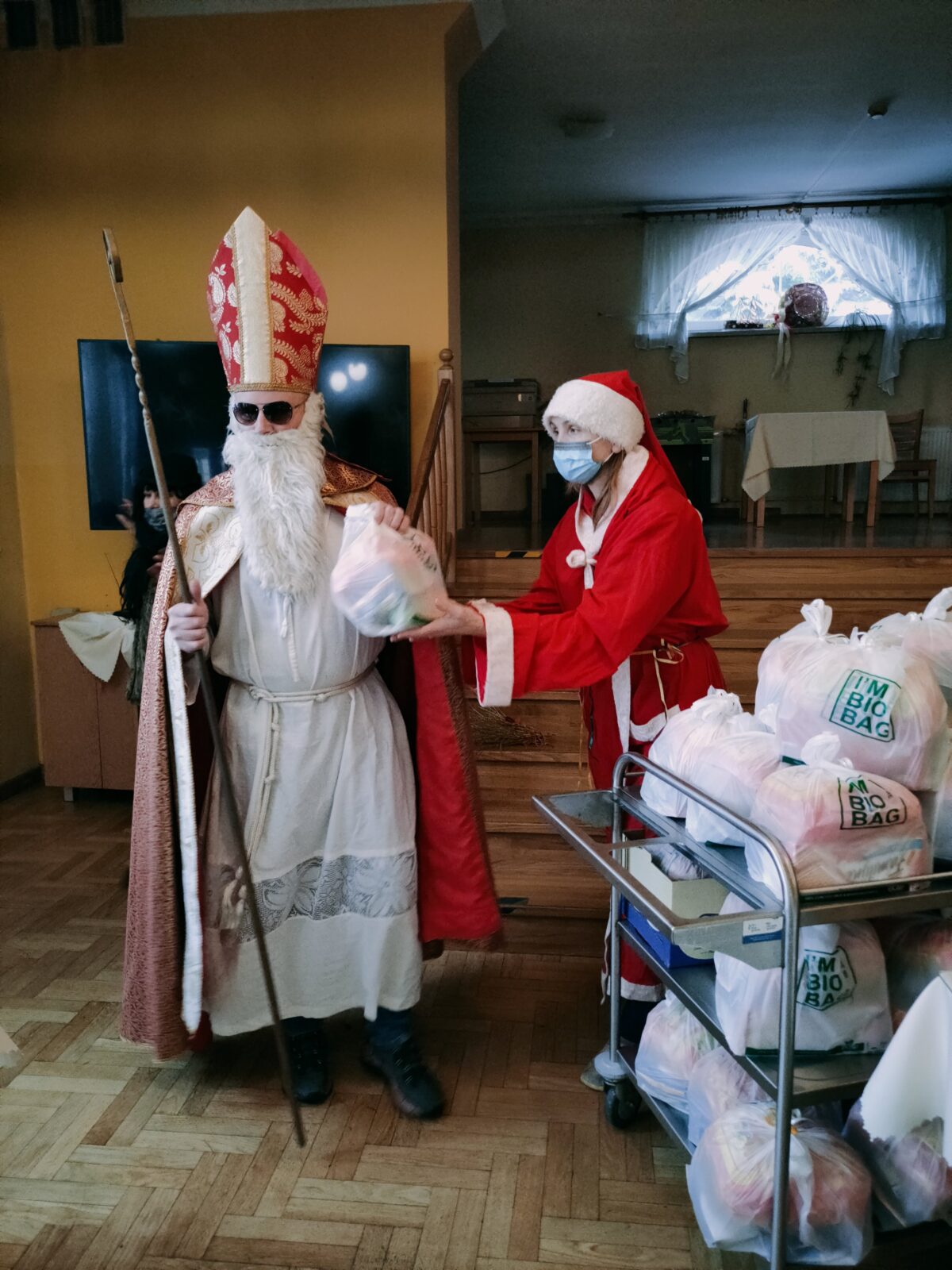 Mężczyzna w stroju biskupa Mikołaja bierze prezent od kobiety w stroju Mikołaja. Obok wózek z prezentami.