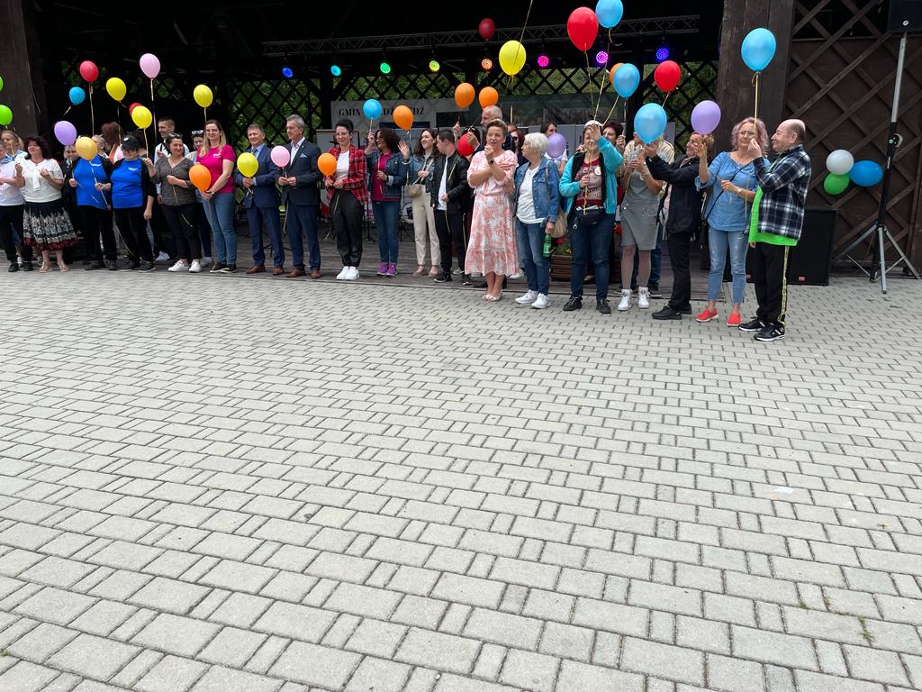grupa osób stojąca przed sceną, na kostce brukowej. Każda osoba trzyma jeden balon. 