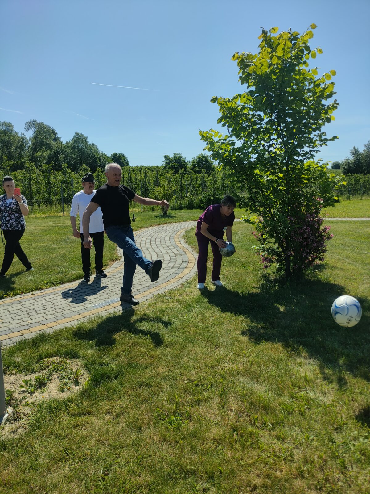 Mężczyzna w pozycji po kopnięciu piłki, w tle dwie osoby - mężczyzna i kobieta, po prawej stronie kobieta podnosi piłkę. W prawym dolnym rogu lecąca piłka. Wszystko dzieje się wśród zieleni na zielonej trawie.
